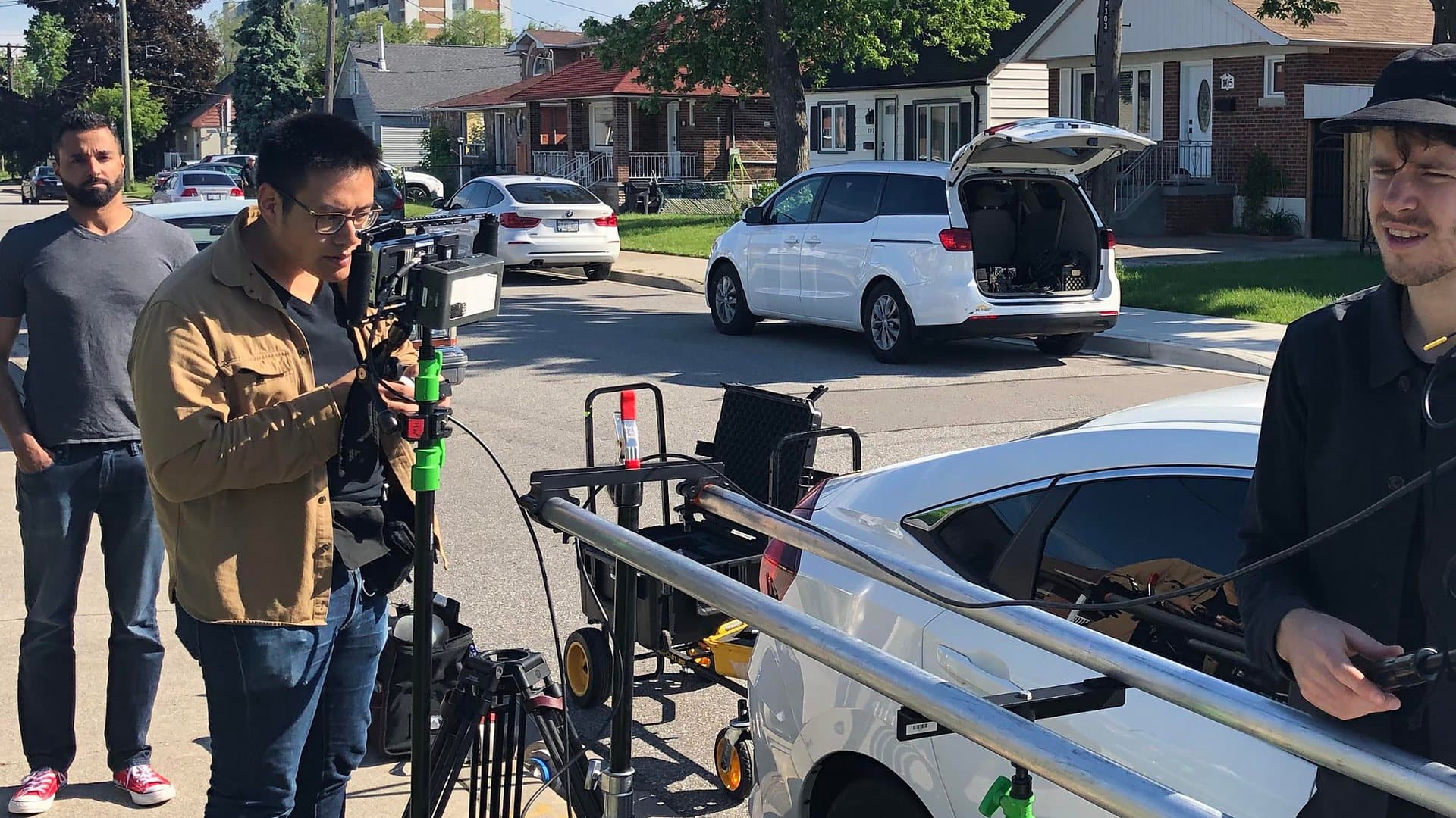 A BGprod crew shooting on a residential street: camera on sticks, a car rig and a grip cart in the road.